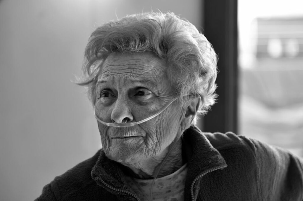 Black and white portrait of an elderly woman with a nasal cannula, expressing resilience and wisdom in her gaze.